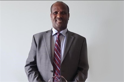 [ai] A man in a suit and tie standing against a plain background, smiling while holding a crystal award in his hands.